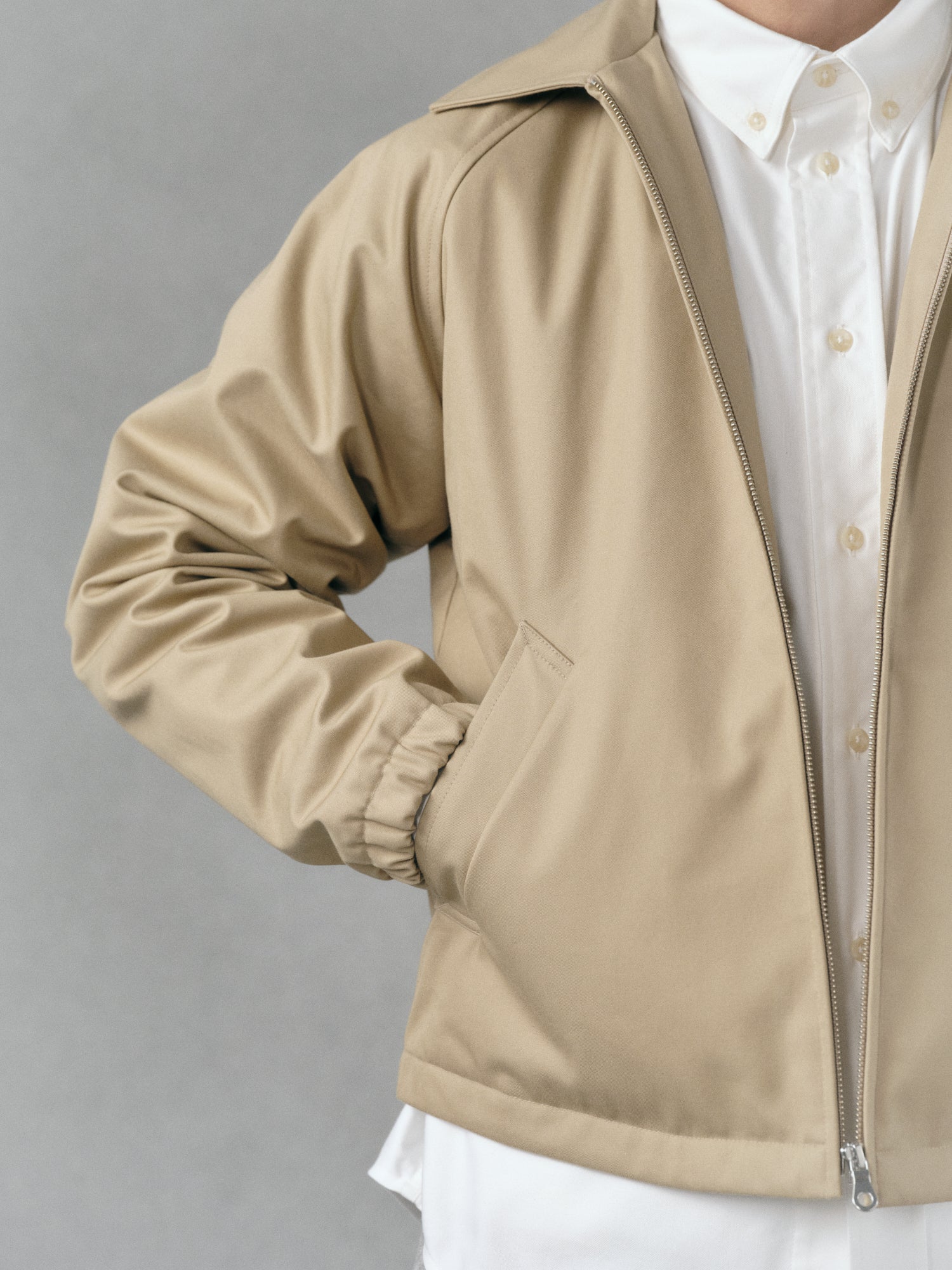 A detail focused photography of a Beige water-repellent made in japan jacket worn over a finely designed white oxford shirt on a gray hand-painted background.
Artisanal approach, Designed in Switzerland, Made in Japan.