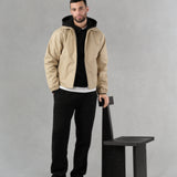 Man wearing a minimal water-repellent beige jacket, a black tailored french-terry hoodie and black straight cut refined jogger pant in french-terry standing next to a designer black wooden chair on a gray artisanal hand-painted background.
