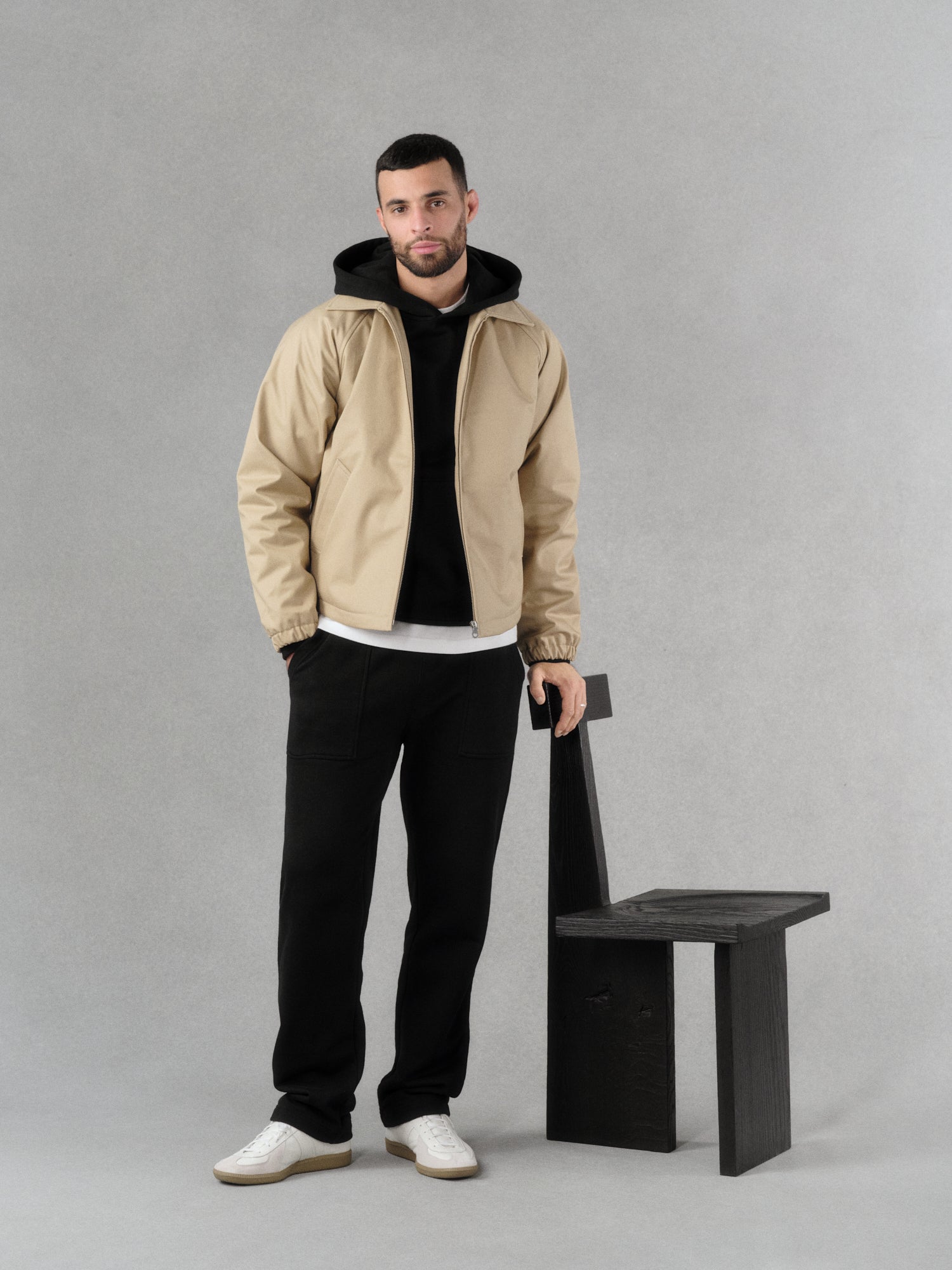 Man wearing a minimal water-repellent beige jacket, a black tailored french-terry hoodie and black straight cut refined jogger pant in french-terry standing next to a designer black wooden chair on a gray artisanal hand-painted background.

